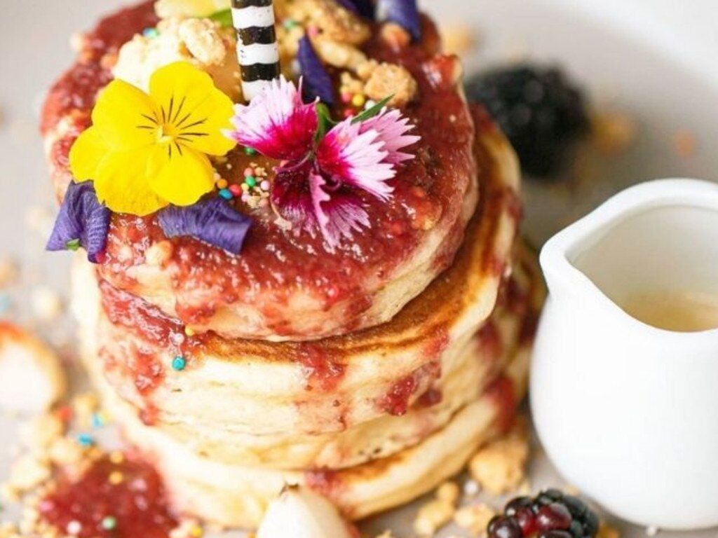 A stack of pancakes topped with edible flowers. A small jug of maple syrup is set to the side.