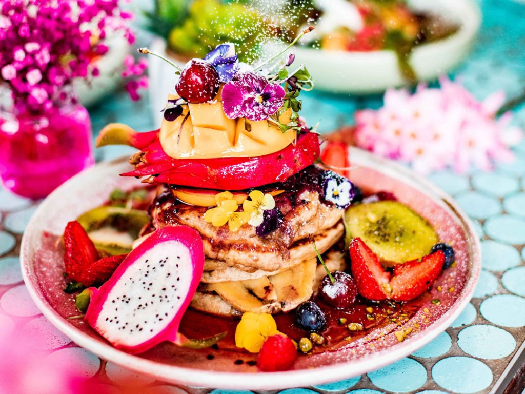 A stack of pancakes with fruit and edible flowers on top and around the plate. The background is artfully out of focus, featuring colourful dishes, flowers and decor.