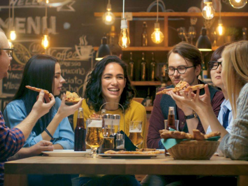 A group of six friends seated around a table, eating pizza and chatting.